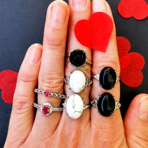 May include: A hand wearing six rings. The rings feature black and white oval stones, a black rose, and a red stone. The rings are on a black background with red hearts.