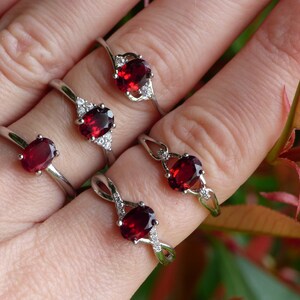 May include: Four silver rings with red oval gemstones and clear accents. The rings are on a hand wearing no other jewelry.