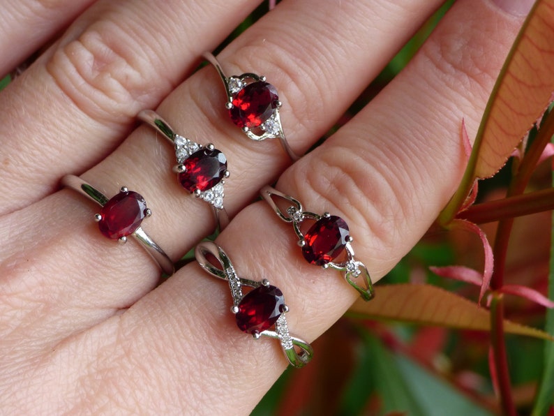 May include: Four silver rings with red oval gemstones and clear accents. The rings are on a hand wearing no other jewelry.
