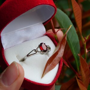 May include: A silver ring with a red oval gemstone and smaller clear gemstones set in a heart-shaped red velvet box.