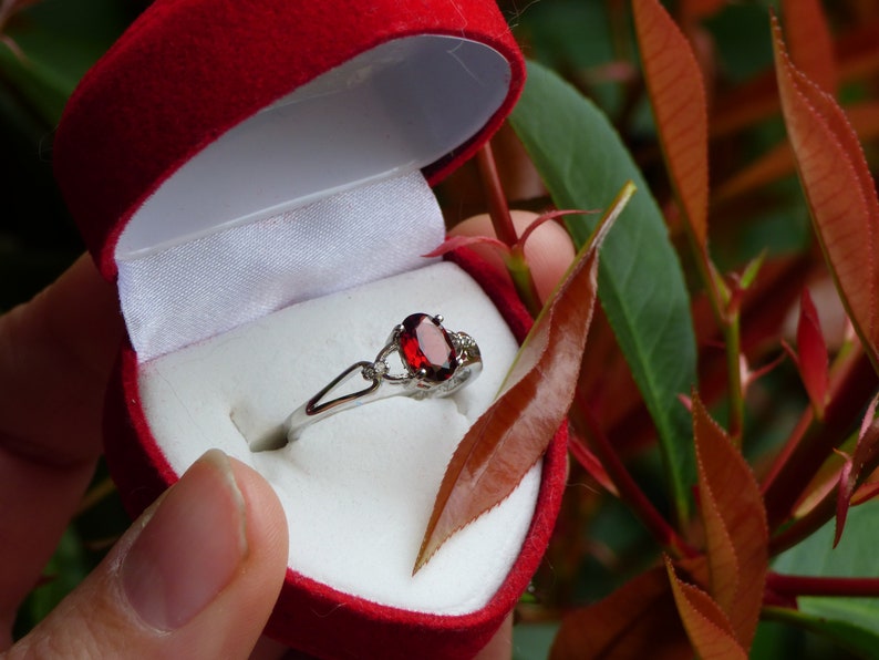 May include: A silver ring with a red oval gemstone and smaller clear gemstones set in a heart-shaped red velvet box.