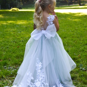 May include: A young girl wearing a light blue tulle dress with a white lace overlay and a large white bow. The dress has a long train with white lace detailing.