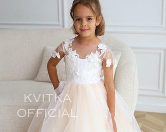 May include: A young girl wearing a white tulle dress with a lace bodice and floral embellishments. The dress has a peach underlay and a full skirt. The girl is standing in front of a white couch.