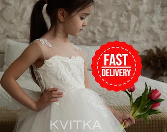 May include: A young girl in a white tulle dress with lace detailing and a sheer bodice. She is holding a bouquet of pink tulips. The dress has a full skirt and is perfect for a special occasion. The text "KVITKA OFFICIAL" is printed on the dress.