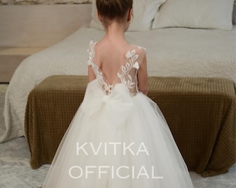May include: A floor-length white dress featuring a sheer illusion back with white floral embroidery, a large tulle bow at the waist, and a full, layered tulle skirt. The text KVITKA OFFICIAL is centered over the skirt of the dress.