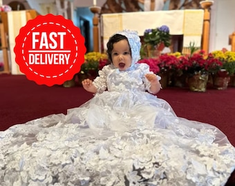 Op de afbeelding: Een witte baby doopjurk met kantdetails en een bijpassende muts. De jurk is erg wijd en heeft een lange sleep. De baby draagt de jurk en de muts en zit op een rood tapijt.