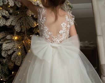 May include: A white sleeveless dress featuring a sheer illusion back with intricate floral embroidery and sequin embellishments. The bodice includes a row of small buttons, and a large tulle bow is attached at the waist above a layered tulle skirt.