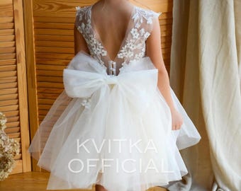 May include: A young girl wearing a white tulle dress with a large bow and floral lace detailing. The dress has a low back and is accented with a white bow. The dress is labeled "Kvitka Official".