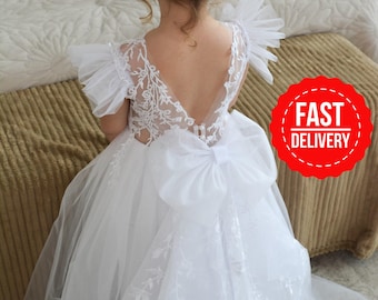 May include: A white floor-length dress featuring a sheer back with intricate floral lace embroidery, ruffled tulle sleeves, and a large tulle bow at the waist. The skirt is made of layered tulle fabric with matching floral lace detailing.