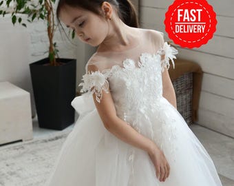May include: A white floor-length dress featuring a sheer illusion neckline, off-the-shoulder sleeves, and intricate floral lace appliques that extend down the bodice. The skirt is made of layered tulle, and a large bow is visible at the back waist.