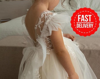 May include: A sleeveless white dress featuring a sheer bodice with intricate floral embroidery, ruffled tulle cap sleeves, and a large tulle bow at the waist. The skirt is made of layered tulle, and the back of the bodice is open with a deep V-shape.