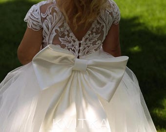 Pode incluir: Um vestido de menina das flores de tule branco com um corpete de renda e um grande laço de cetim branco nas costas. O vestido tem mangas curtas e um decote em V.