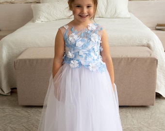 Puede incluir: Un vestido blanco largo hasta el suelo con un corpiño sin mangas que presenta patrones de hojas bordadas en azul claro y aplicaciones florales blancas en tres dimensiones. La falda está hecha de tul en capas, creando una silueta amplia.