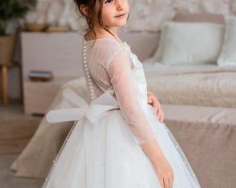 May include: A white floor-length dress featuring sheer long sleeves, a row of small buttons along the back, and a large tulle bow at the waist. The skirt is made of layered tulle fabric embellished with scattered small white pearls throughout.