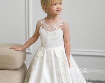 May include: A young girl wearing a white formal dress with floral lace detailing on the bodice and sheer neckline. The dress has a full skirt and the text "KVITKA OFFICIAL" is visible.
