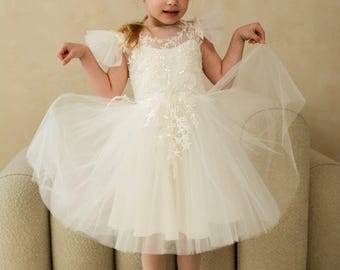 May include: An ivory-colored dress featuring a sheer illusion neckline, delicate floral lace embroidery with subtle sequin embellishments on the bodice, fluttery tulle sleeves, and a voluminous, multi-layered tulle skirt that reaches the knees.