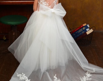 May include: A floor-length white dress featuring a sheer illusion back with delicate white floral lace embroidery, a large tulle bow at the waist, and a voluminous layered tulle skirt that extends into a long train with matching floral lace trim.