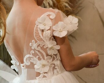 Puede incluir: Un vestido blanco sin mangas con espalda transparente de ilusión, bordados florales blancos intrincados y flores de tela tridimensionales acentuadas con pequeñas perlas centrales. El corpiño se une a una falda de tul con varias capas.