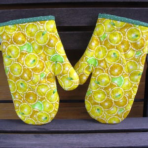 Könnte beinhalten: Ein Paar gelbe und grüne Ofenhandschuhe mit einem Zitronen- und Limettenmuster. Die Handschuhe sind aus Stoff und haben eine gesteppte Textur.