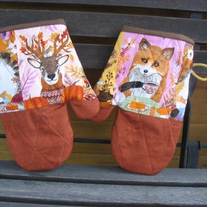 Könnte beinhalten: Zwei braune, gesteppte Ofenhandschuhe mit einem rosa-orangenen Stoffdesign. Ein Handschuh zeigt ein Reh mit Geweih, der andere einen Fuchs, der eine Tasse hält. Beide Handschuhe haben eine braune, gesteppte Rückseite.