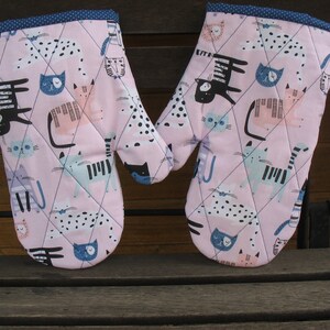 Könnte beinhalten: Ein Paar rosa Ofenhandschuhe mit gestepptem Design und blauem Besatz. Die Handschuhe zeigen ein sich wiederholendes Muster aus schwarzen, weißen und blauen Katzen.