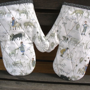 Könnte beinhalten: Ein Paar weiße Topflappen mit gestepptem Design, das ein sich wiederholendes Muster aus Schafen, Kindern und Blumen zeigt. Die Topflappen haben einen grauen und weißen Saum.