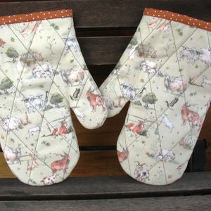 Könnte beinhalten: Ein Paar gesteppte Ofenhandschuhe mit Bauernhoftierdruck. Die Handschuhe haben einen hellgrünen Hintergrund mit einem Muster aus Schafen, Ziegen und anderen Nutztieren. Die Ränder sind mit braunem Stoff mit weißen Punkten verziert.