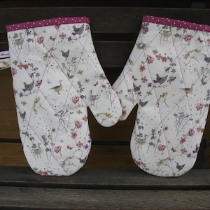Könnte beinhalten: Ein Paar weiße Ofenhandschuhe mit einem gesteppten Muster mit Hühnern, Gänsen, Schmetterlingen und floralen Elementen. Die Handschuhe haben eine rosa gepunktete Bordüre und eine Schlaufe zum Aufhängen.