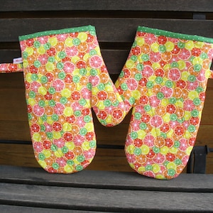 Könnte beinhalten: Ein Paar Ofenhandschuhe mit einem Zitrusfruchtmuster. Die Handschuhe sind gelb, orange und grün mit weißem Hintergrund. Die Handschuhe haben einen grünen Saum und weiße Punkte.