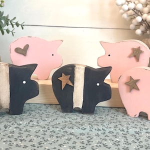 May include: Wooden pig figurines in pink and black with heart and star accents. The pigs have a rustic, farmhouse style. Some have a white stripe down the middle. The figurines are displayed on a wooden base.