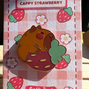 Puede incluir: Un pin de esmalte metálico que muestra un capibara marrón comiendo una fresa roja con hojas verdes. El pin está sujeto a una tarjeta de soporte rectangular con un patrón de cuadros vichy rosa y blanco, ilustraciones de fresas y el texto CAPPY STRAWBERRY.