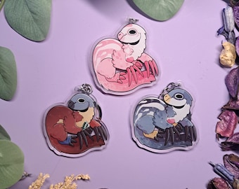 May include: Three acrylic keychains featuring cartoon-style theropod dinosaurs in pink, blue, and red. Each keychain has a silver keyring and a clear border. The dinosaurs are in a curled-up pose on a purple background.