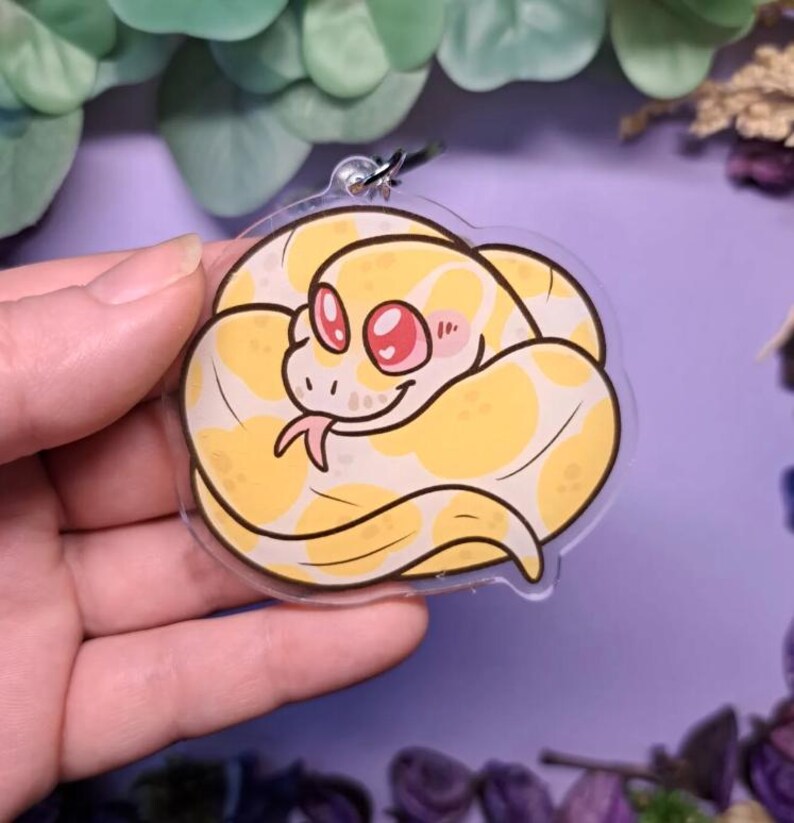 Op de afbeelding: Een heldere acryl bedel met een cartoon slang in een opgerolde positie. De slang is geel met witte vlekken, roze ogen en een roze blos. De bedel heeft een zilverkleurige sluiting.