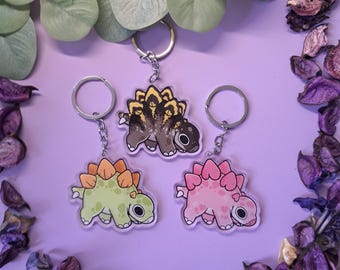 Könnte beinhalten: Drei Schlüsselanhänger in Dinosaurierform in Grün, Rosa und Braun mit orangefarbenen, rosa und gelben Akzenten. Jeder Schlüsselanhänger hat einen silbernen Schlüsselring. Die Schlüsselanhänger liegen auf einer lila Oberfläche mit dekorativen Blättern und Trockenblumen.