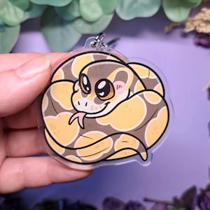 Op de afbeelding: Een heldere acryl sleutelhanger met een cartoon slang opgerold. De slang heeft een bruin en geel patroon, met grote ogen, een roze tong en roze wangen. Een zilverkleurige metalen ring is aan de bovenkant bevestigd.