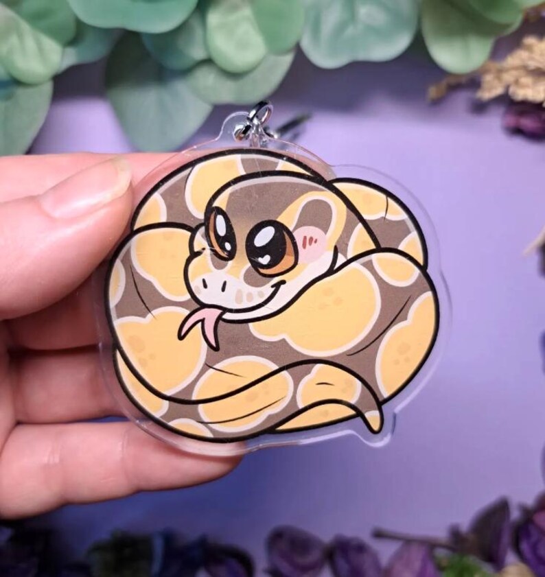 Op de afbeelding: Een heldere acryl sleutelhanger met een cartoon slang opgerold. De slang heeft een bruin en geel patroon, met grote ogen, een roze tong en roze wangen. Een zilverkleurige metalen ring is aan de bovenkant bevestigd.