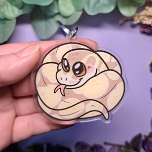 Op de afbeelding: Een heldere, ronde sleutelhanger met een cartoon slang met een cr&egrave;me- en bruin patroon. De slang heeft grote, vriendelijke ogen, een roze tong en roze wangen. De sleutelhanger wordt omhoog gehouden door een zilveren metalen ring.