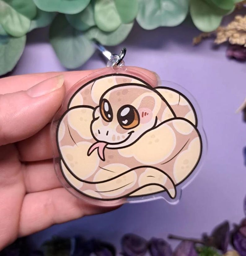 Op de afbeelding: Een heldere, ronde sleutelhanger met een cartoon slang met een cr&egrave;me- en bruin patroon. De slang heeft grote, vriendelijke ogen, een roze tong en roze wangen. De sleutelhanger wordt omhoog gehouden door een zilveren metalen ring.