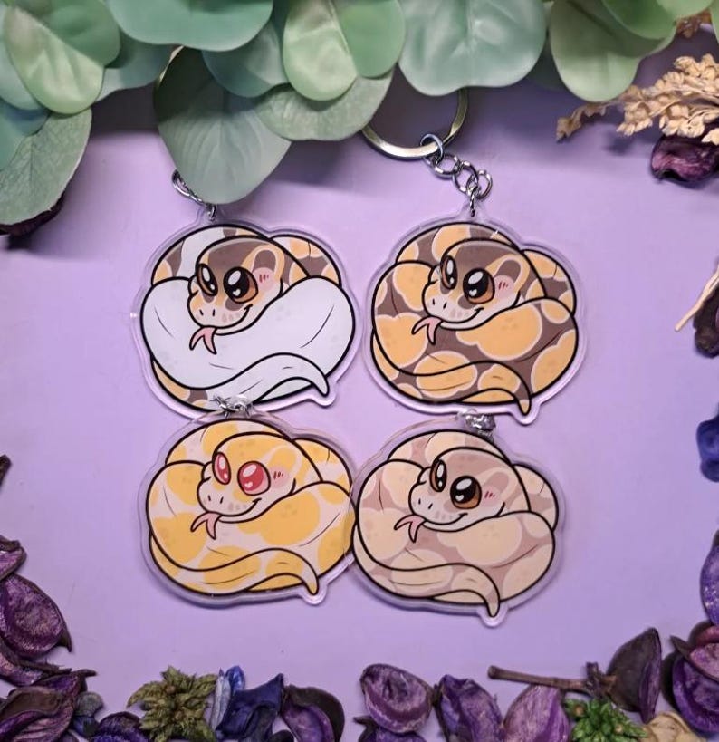 Op de afbeelding: Vier acryl sleutelhangers met cartoon slangen in verschillende kleuren. De slangen zijn opgerold en hebben schattige, expressieve gezichten. De sleutelhangers hebben zilverkleurige metalen ringen. De achtergrond is zacht paars met decoratief blad.