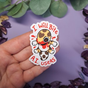 Puede incluir: Una pegatina blanca con un diseño de criatura de dibujos animados. La criatura es amarilla y marrón con un patrón de corazón, acentos rojos como sangre y el texto "I Will Bite A.I. Users".
