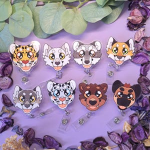 May include: A collection of eight animal-themed retractable badge holders. Each badge features a different cartoon animal face, including a leopard, wolf, raccoon, fox, snow leopard, bear, and dog. The badges are clear with a plastic clip and a retractable cord.