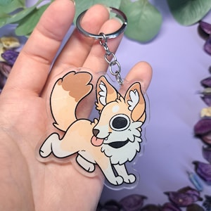May include: A clear acrylic keychain featuring a cartoon fox with a light orange and white color scheme. The fox has a happy expression with its tongue sticking out. The keychain includes a silver metal ring and chain.