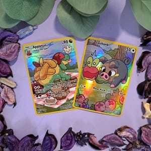 Puede incluir: Dos cartas coleccionables de Pokémon con lámina holográfica. Una carta presenta a Appletun con los textos "Melting Sweetness" y "Wild Tackle". La otra carta presenta a Lechonk con el texto "Rear Kick". Ambas cartas tienen bordes amarillos.