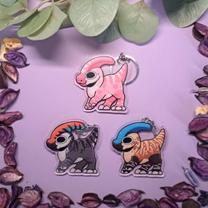 May include: Three dinosaur keyrings in a cartoon style. One is pink with heart patterns, another is grey with orange and blue accents, and the third is tan with a blue crest. All have silver key rings on a purple background.