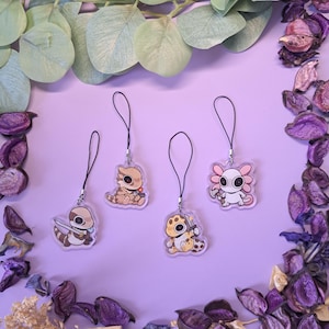 May include: Four acrylic charms with black lanyards, featuring cartoon reptile and amphibian designs. The charms include a brown and white dinosaur with a sword, a tan and brown dinosaur, a yellow and brown dinosaur with a knife, and a pink axolotl with a hammer.