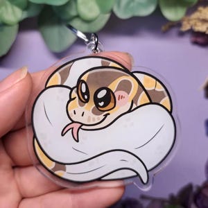 Op de afbeelding: Een heldere acryl sleutelhanger met een cartoon slang opgerold in een bal. De slang heeft een wit lichaam met bruine en gele markeringen, grote ogen en een roze tong. Een zilveren sleutelring is aan de bovenkant bevestigd.