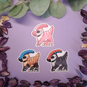 May include: Three cartoon dinosaur stickers. One is pink with heart patterns, another is brown with a blue crest, and the third is grey with a red and blue crest. The stickers are on a purple background, surrounded by decorative elements.