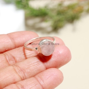 May include: A silver ring featuring a round, light pink rose quartz gemstone. The ring has a simple band and a bezel setting. The text "Natural Gems Glare" is visible on the ring. The ring is shown on a person's finger.