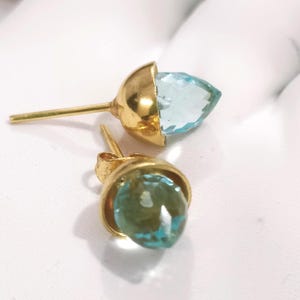 May include: A pair of gold-colored stud earrings. Each earring features a light blue, teardrop-shaped gemstone set within a gold bezel. The earrings are displayed on a white surface.