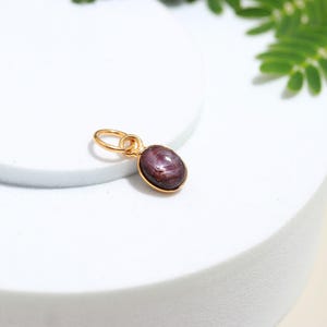 May include: A gold pendant featuring a dark red oval gemstone. The gemstone is set in a gold bezel and attached to a small gold loop. The pendant is shown on a white surface.